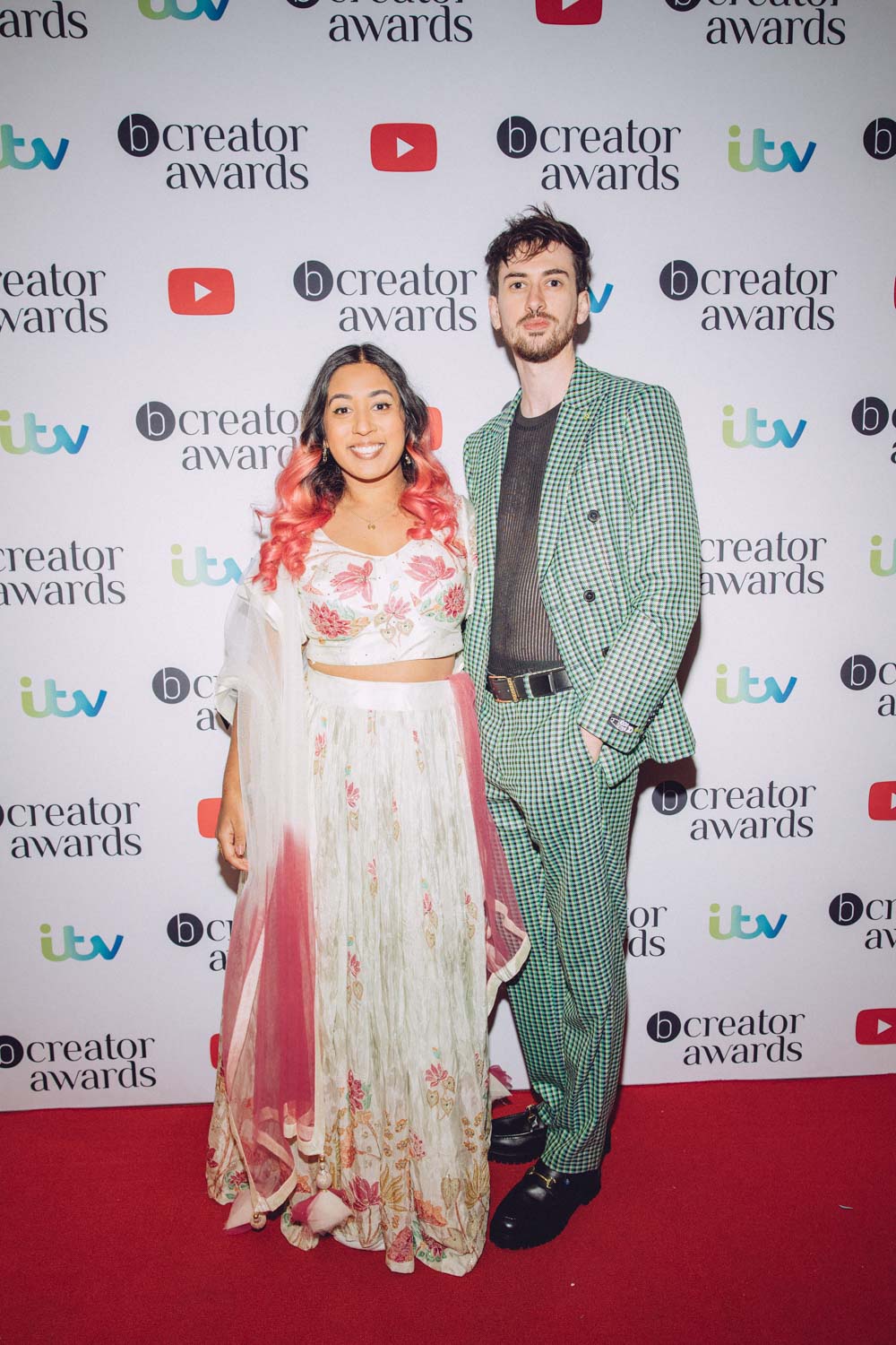 Shaaba wearing a pink and white floral gown accessorised with a shawl and pink hair highlights. Jamie wearing a checked black and green suit from TWISTED TAILOR.